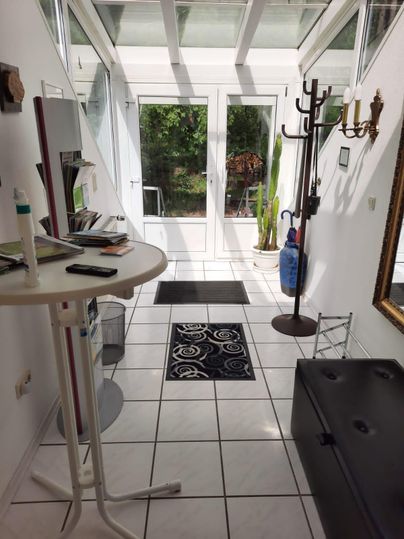 Eine helle Diele mit einem runden Tisch, einem Teppich und einem Garderobenständer. Durch die Glastüren ist ein Gartenbereich sichtbar.