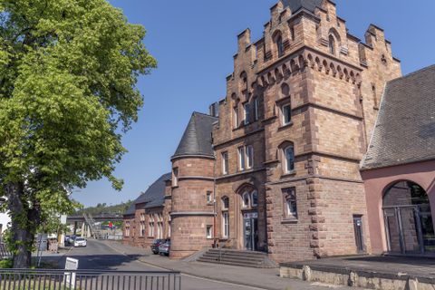 Ein historisches Gebäude im neugotischen Stil mit Türmen und roten Ziegeln. Im Hintergrund sind Bäume und ein klarer Himmel zu sehen.
