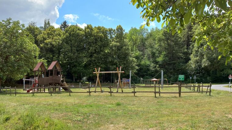 Spielplatz mit Holzhäuschen, Schaukeln und Klettergerüst, umgeben von Bäumen und Wiese, unter blauem Himmel mit Wolken.