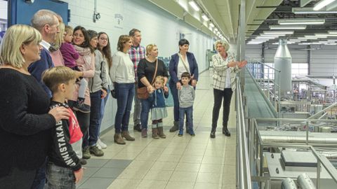 Eine Gruppe von Menschen, darunter Kinder, bei einer Führung in einer Fabrikhalle. Eine Frau erklärt etwas, während die Gruppe zuhört.