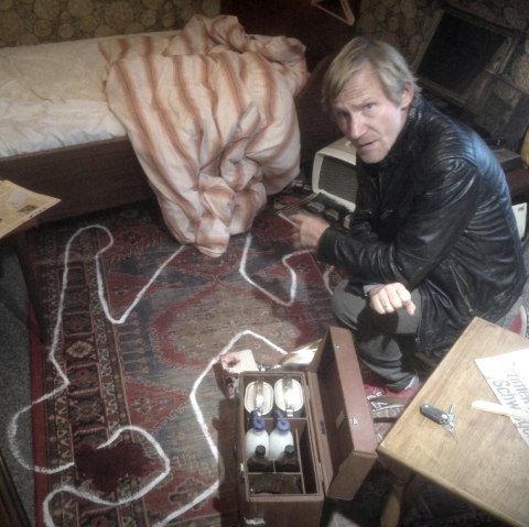 A man is kneeling on the floor in a bedroom. The room is untidy and the outline of a corpse is drawn in chalk on the carpet in front of him.