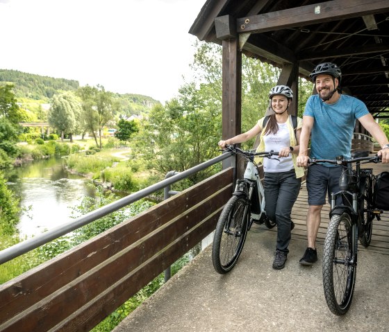 Two people with bicycles are standing on a covered wooden bridge in the Gerolstein spa gardens. A river and green landscape can be seen in the background., © Eifel Tourismus GmbH, Dominik Ketz