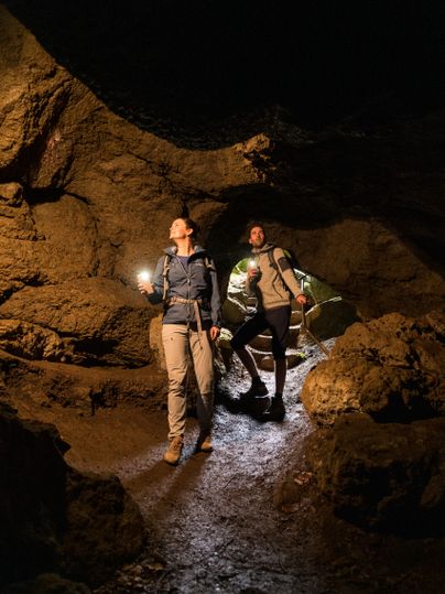 Two people enter a dark cave. Both are shining the torches on their mobile phones. The rock walls of the cave surround the hikers.