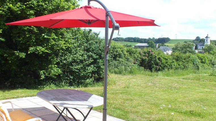 Een rode parasol staat naast een tafel en stoel op een terras. Op de achtergrond zijn groene weiden en bomen te zien.