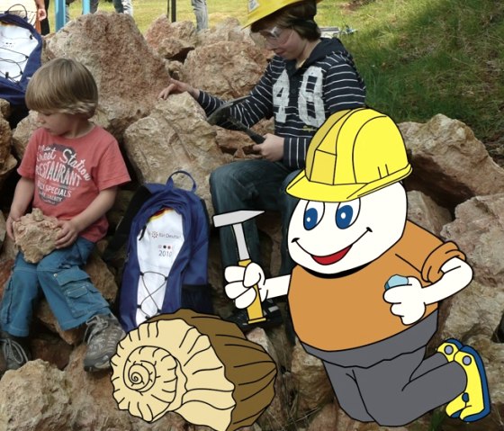 Cartoonfigur Willi Basalt mit grünem Shirt, grauer Hose, gelben Schuhen und gelben Helm kniet vor einem Fossil und hat einen Hammer in der Hand. Hinter ihm ein Foto, auf dem Kinder weitere Gesteine auf dem Geoacker Gerolstein untersuchen.
