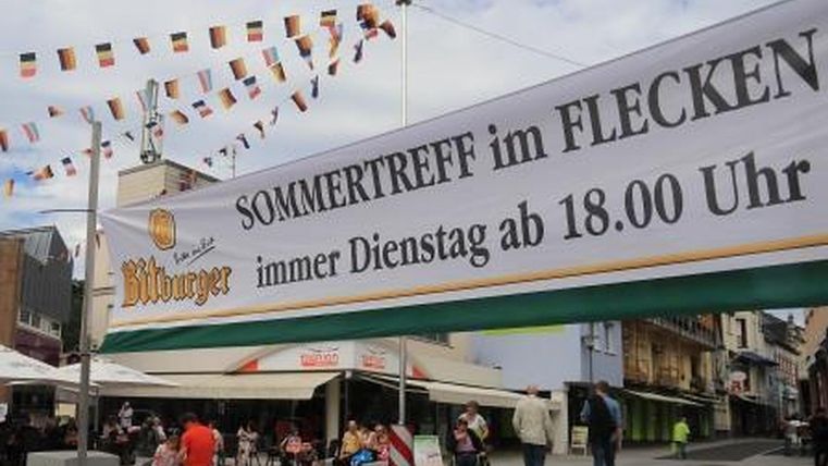 Eine belebte Stadtfläche mit einem großen Banner, das auf ein Sommerereignis hinweist. Im Hintergrund sind Menschen und Terrassen von Cafés zu sehen.