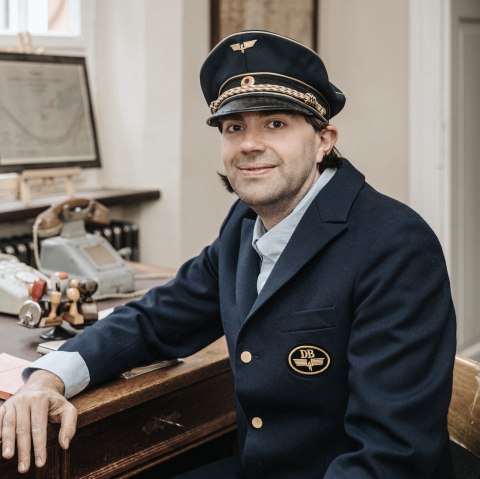 Mann in historischer Schaffnerkleidung sitzt an einem Schreibtisch, auf dem eine Schreibmaschine, Stempel und ein antikes Telefon stehen und schaut in die Kamera.