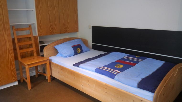 Ein einfaches Schlafzimmer mit einem Holzbett und einem Stuhl. Die Bettwäsche ist blau und bunt; die Wände sind hell.