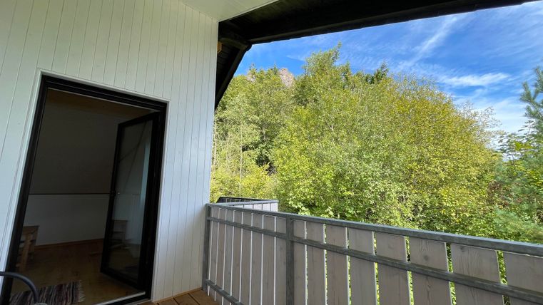 Un balcon avec un sol en bois et une porte ouverte. La vue montre un feuillage vert luxuriant et un ciel bleu.