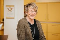 Eine Frau steht hinter einem Tresen eines Postschalters. Hinter ihr Schränke und das Posthorn-Logo.