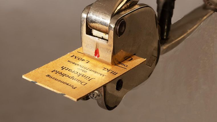 Eine silberne Lochzange stanzt ein Stück Papier mit Text. Die Zange hat einen roten Markierungspunkt.