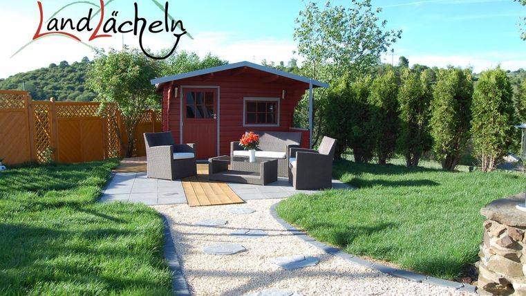 Ein gemütlicher Garten mit einem roten Holzhaus und einem Sitzbereich. Der Rasen ist grün und gut gepflegt, umgeben von Bäumen und einem Zaun.