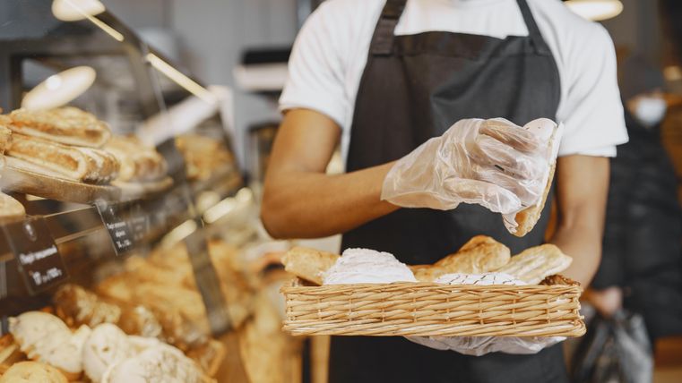 Ein Bäcker mit Schürze und Handschuhen legt frisches Gebäck in einen Korb in einer Bäckerei.