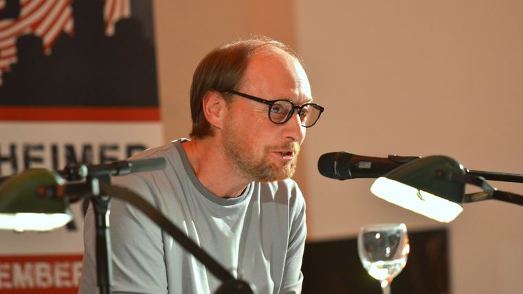 Ein man sitzt an einem Tisch und spricht in ein Mikrophon.