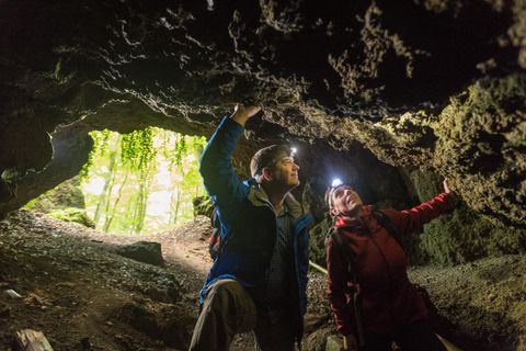 Zwei Personen mit Strinlampen erkunden die Felswände einer Höhle. Im Hintergrund ist der Höhlenausgang.