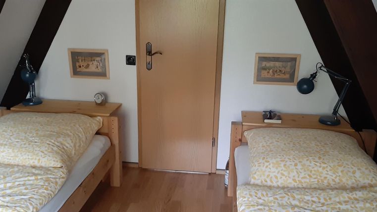 Ein gemütliches Schlafzimmer mit zwei Einzelbetten und warmen Holzböden. An den Wänden hängen Bilder und eine Tür führt nach draußen.