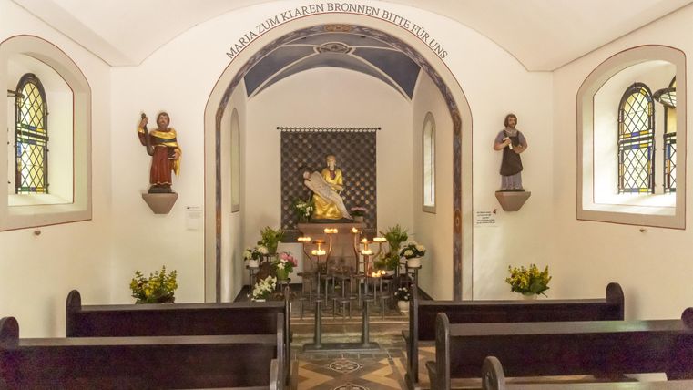 Innenansicht der Büschkapelle mit Bänken, Statuen und einem Altar. Kerzen brennen vor einer goldenen Statue. Schriftzug: 'Maria zum klaren Brunnen bitte für uns'.