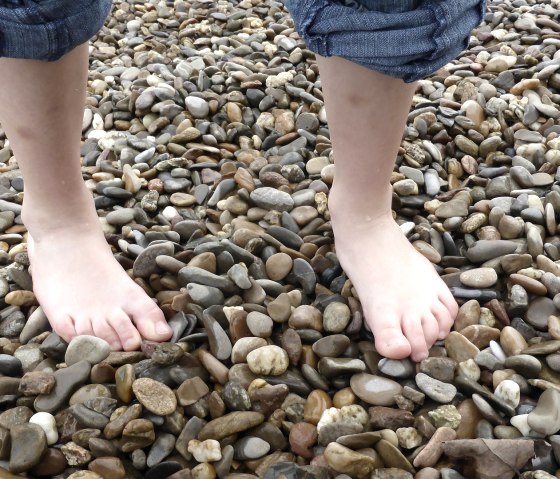 Two bare feet are standing on a pebble beach. The person is wearing rolled-up jeans., © pixabay
