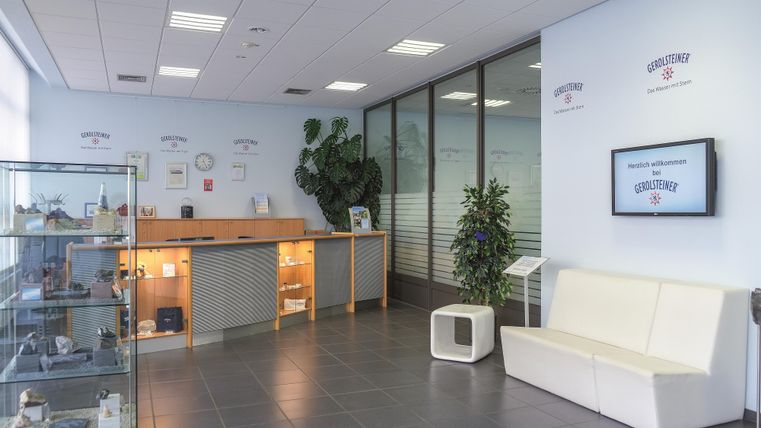 Modernes Besucherzentrum mit Empfangstresen, Glasvitrine, weißen Sitzbänken und Pflanzen. An den Wänden sind Gerolsteiner-Logos sichtbar.