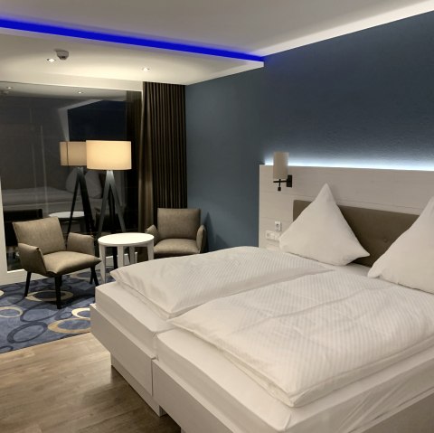 Modern eingerichtetes Hotelzimmer im Seehotel am Stausee mit einem gro&szlig;en wei&szlig; bezogenen Doppelbett, LED-Licht, Teppich und einem Beistelltisch mit St&uuml;hlen.