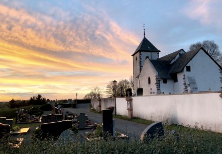 Eine Wehrkirche bei Sonnenuntergang, umgeben von einem Friedhof. Der Himmel ist in warmen Orange- und Blaut&ouml;nen gef&auml;rbt.