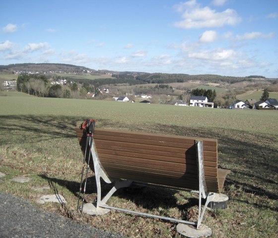 A bench with walking sticks stands in front of a wide, green landscape with hills and scattered houses under a blue sky., © Touristik GmbH Gerolsteiner Land, Ute Klinkhammer