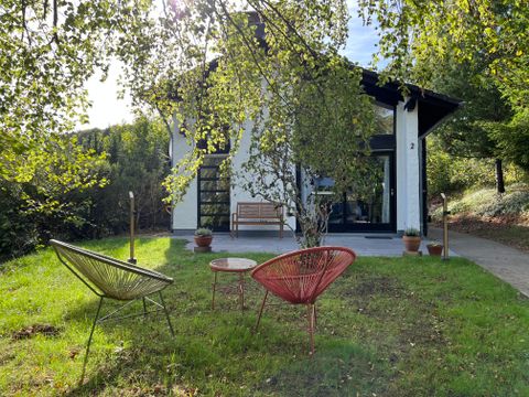 Een modern huis omgeven door bomen en groen gras. Voor het huis staan twee stijlvolle stoelen en een kleine tafel.