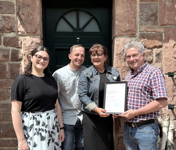 Vier gelukkige en lachende mensen staan voor een toegangsdeur en houden trots het certificaat voor de classificatie in hun handen.