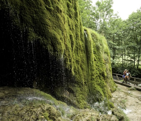 Der Nohner Wasserfall mit moosbewachsenen Felsen. Zwei Personen stehen auf einem Steg im Grünen. Ein idyllischer Zwischenstopp am Kalkeifel-Radweg., © Rheinland-Pfalz Tourismus GmbH/D. Ketz