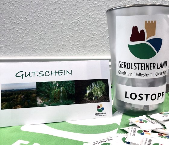 Ein Lostopf mit dem Logo der Ferienregion steht auf einem Tisch, daneben der zu gewinnende Reisegutschein und davor einige Traubenzucker.