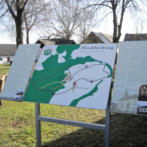 Infotafel mit Karte des Pferdsheckwegs und Texten. Sie steht im Freien, umgeben von Bäumen und Gebäuden. Der Himmel ist klar und sonnig., © Touristik GmbH Gerolsteiner Land, Ute Klinkhammer