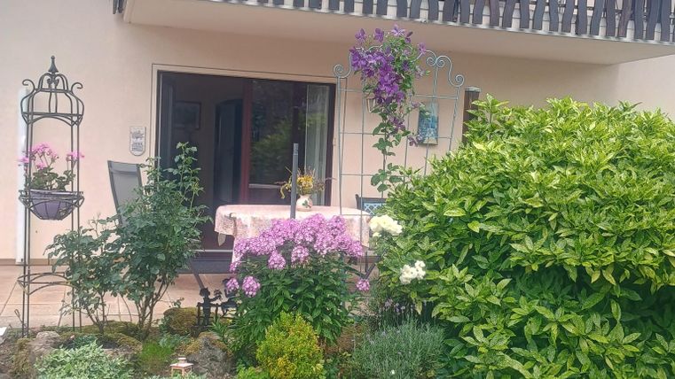 Ein einladendes Haus mit einem Balkon, geschmückt mit blühenden Pflanzen. Der Garten ist bunt und grün, mit abwechslungsreicher Vegetation und einem kleinen Sitztisch.