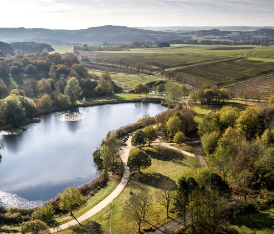 Luchtfoto van een meer in de vallei van Bolsdorf, omringd door herfstbomen en velden, met een pad langs de oever., © Eifel Tourismus GmbH, Dominik Ketz