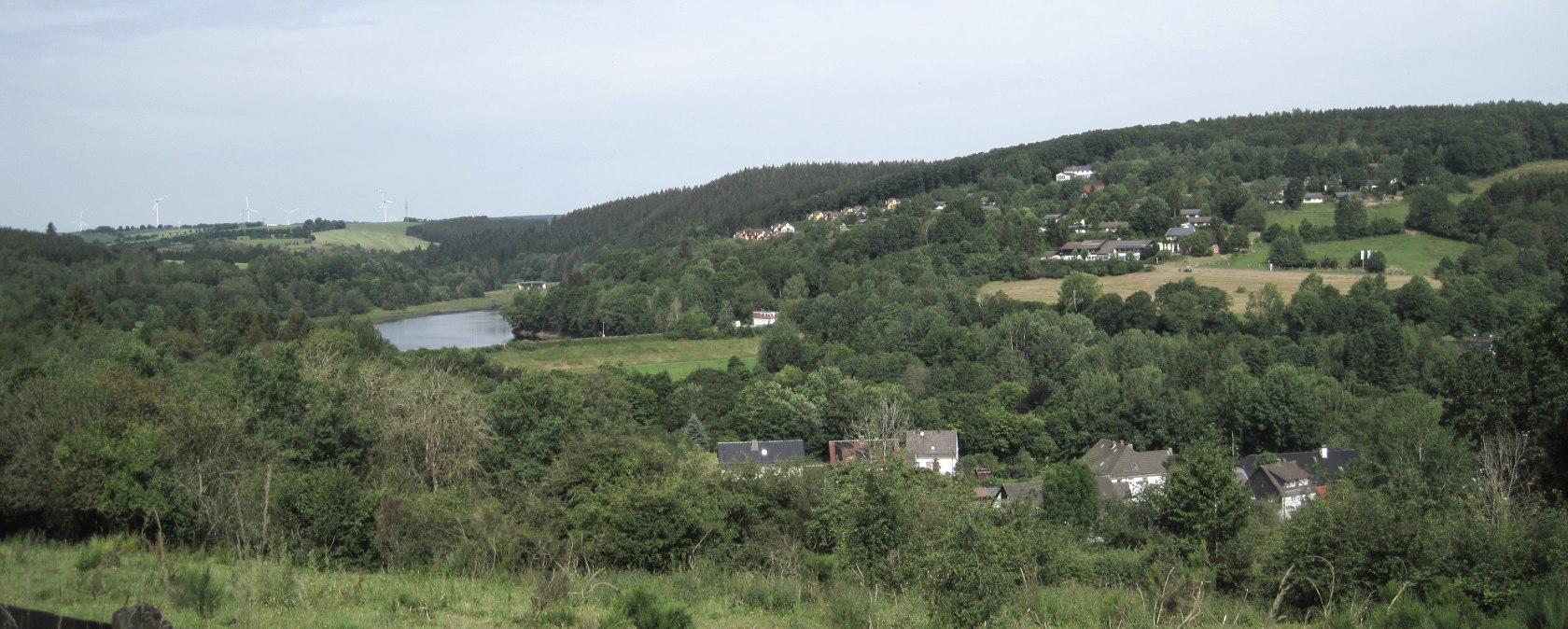 View of Lake Kronenburg, surrounded by green forests and meadows, with wind turbines in the background., &copy; Touristik GmbH Gerolsteiner Land