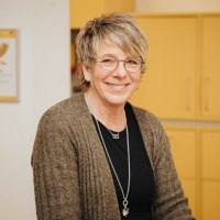 Eine Frau steht hinter einem Tresen eines Postschalters. Hinter ihr Schränke und das Posthorn-Logo.