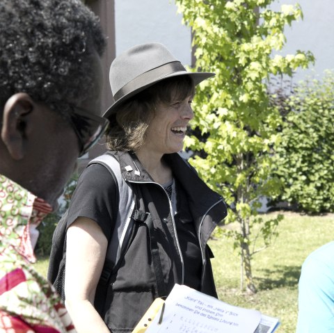 The focus is on a darkly dressed woman in a hat who is laughing a lot. There are other people around her in front of a building.