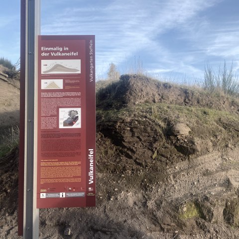 Informationstafel der Vulkaneifel zum Thema "Einmalig in der Vulkaneifel" mit mehrsprachigen Texten und anschaulichen Grafiken. Daneben vulkanische Erdschichten.
