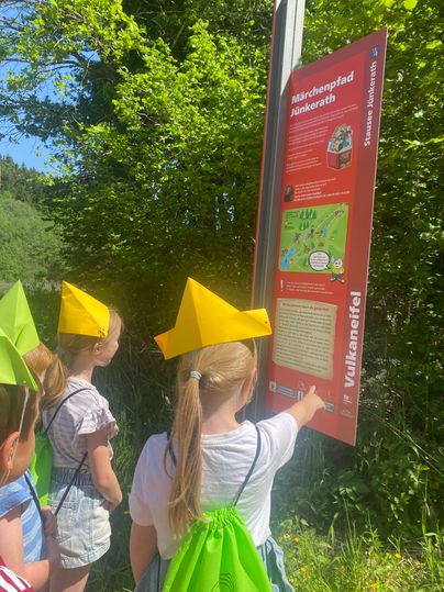 Kinder mit bunten Hüten und grünen Rucksäcken stehen vor einem Schild des Märchenpfads Jünkerath.