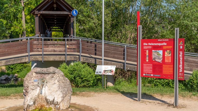 Eine überdachte Holzbrücke und ein Informationsschild zur Helenenquelle im Kyllpark Gerolstein.
