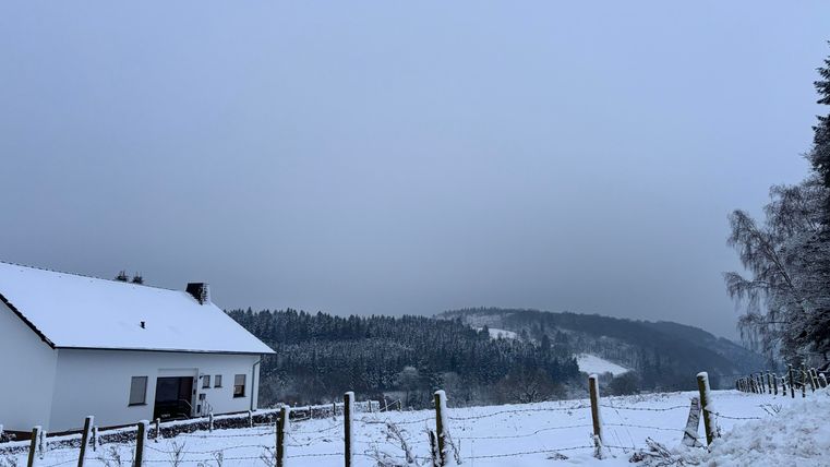 Ein schneebedecktes Landschaftsbild mit einem weißen Haus im Vordergrund. Im Hintergrund sind Bäume und ein bewölkter Himmel zu sehen.