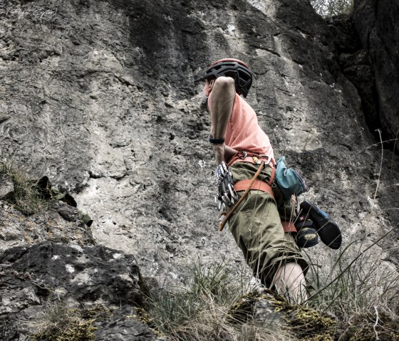 Eine Person mit Helm und Ausrüstung bekleidet steht vor einer steilen Felswand, stützt die Hände in die Hüften und schaut hoch.