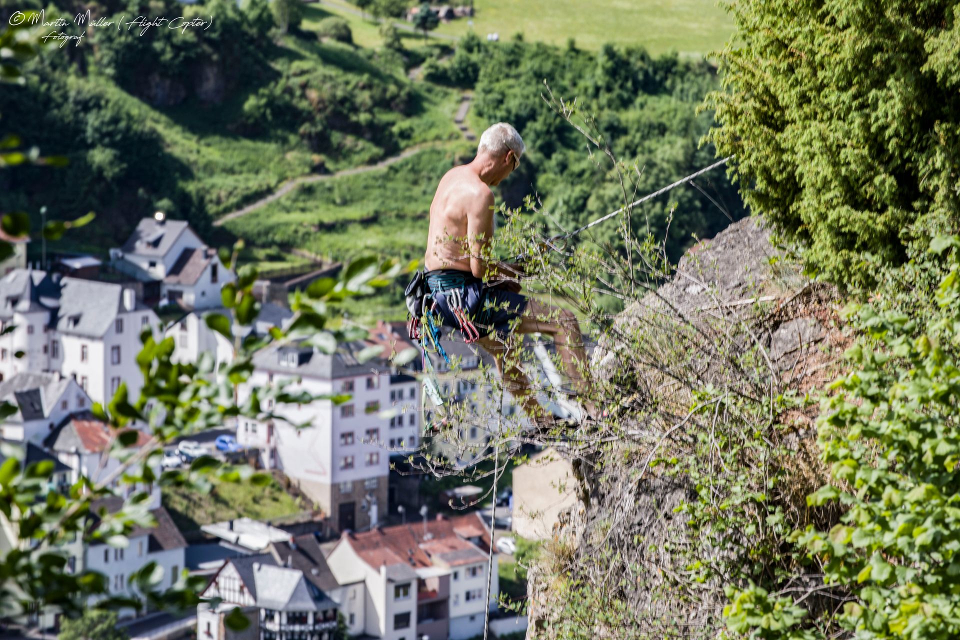 Ein Mann klettert an einem Felsen, gesichert mit Seilen. Im Hintergrund ist ein Dorf mit weißen Häusern und grüner Landschaft zu sehen.