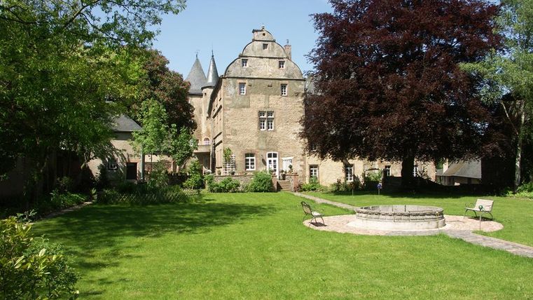 Burg Lissingen mit gepflegtem Garten, Brunnen und Bänken. Historisches Gebäude mit Türmen und Bäumen im Hintergrund.