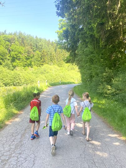 Vier Kinder mit grünen Rucksäcken gehen auf einem Asphaltierten Weg spazieren.