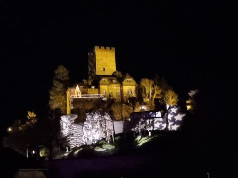 Eine beleuchtete Burg steht auf einem Hügel in der Nacht. Umgeben von Bäumen strahlt sie eine festliche Atmosphäre aus.