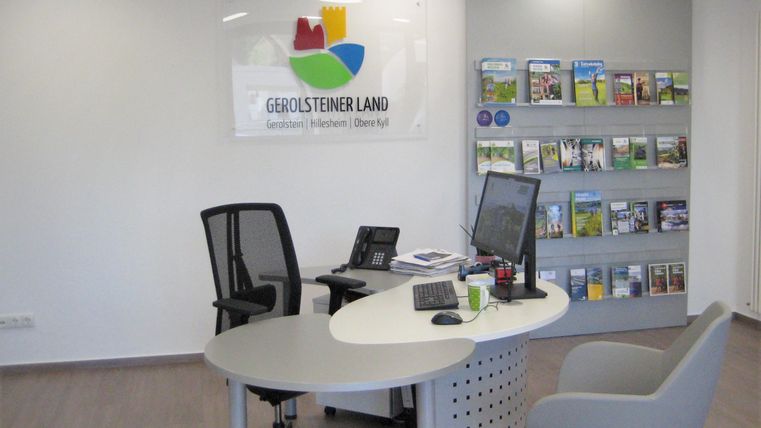 Ein moderner Empfangsbereich mit einem runden Tisch und einem Computer. Im Hintergrund stehen Informationsbroschüren in einem Regal.