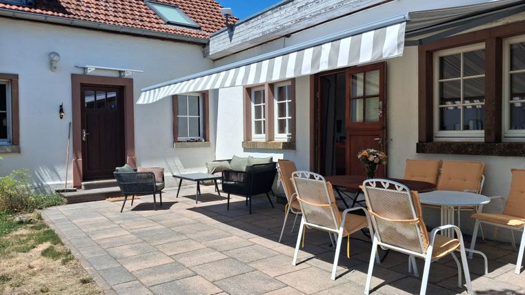 Ein gemütlicher Außenbereich mit einem gepflasterten Boden und mehreren Sitzgelegenheiten. Der Sonnenschirm spendet Schatten vor einem hellen Haus mit einer einladenden Tür.