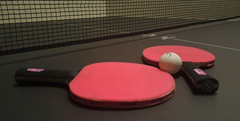 Zwei Tischtennisschläger mit roter Schlagfläche und ein Ball  liegen auf einer dunklen Tischtennisplatte.