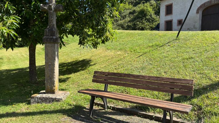 A wooden bench stands next to a stone cross in a meadow. A building with a white façade and dark roof can be seen in the background.
