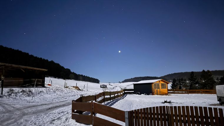 Eine verschneite Landschaft unter dem klaren Nacht Himmel. Ein kleines Holzhaus und ein Zaun sind im Vordergrund sichtbar.